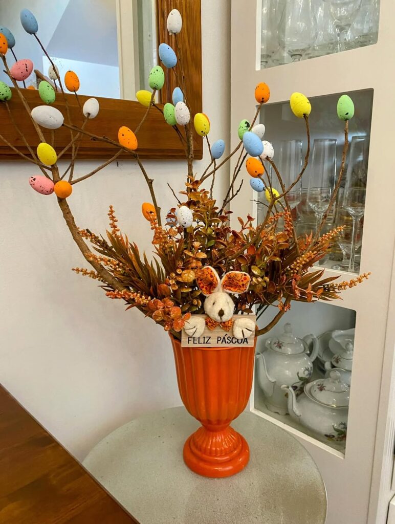 Árvore de Páscoa com galhos secos, ovinhos coloridos e coelho de pelúcia em vaso de cerâmica laranja