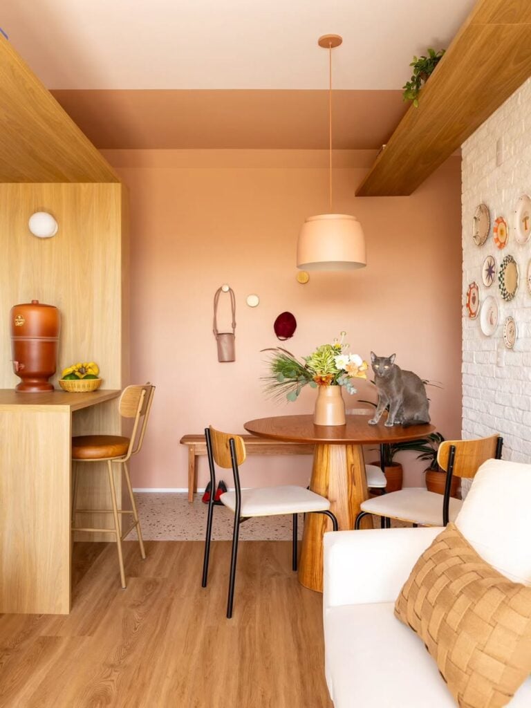 Sala de jantar com mesa redonda de madeira com um vaso de flores e um gato cinza, pendente acima e parede em tom de pêssego