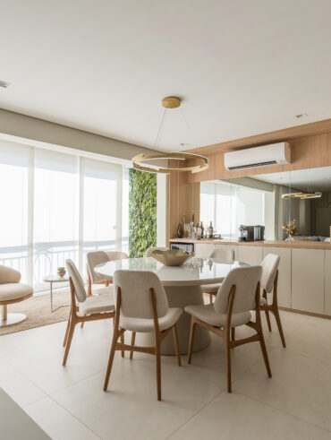 Sala de jantar com mesa retonda, cadeiras em estrutura de madeira com estofados claros, armários claros e espelho na extensão da parede