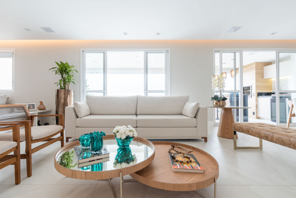 Sala com sofá branco de linhas retas, móveis em madeira e mesas de centro com livros e vasos decorativos