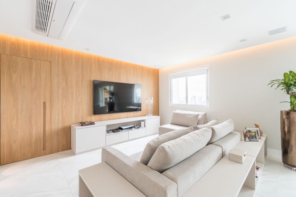 Sala com parede branca e painel amadeirado atrás da tv, rack e sofás em cores claras
