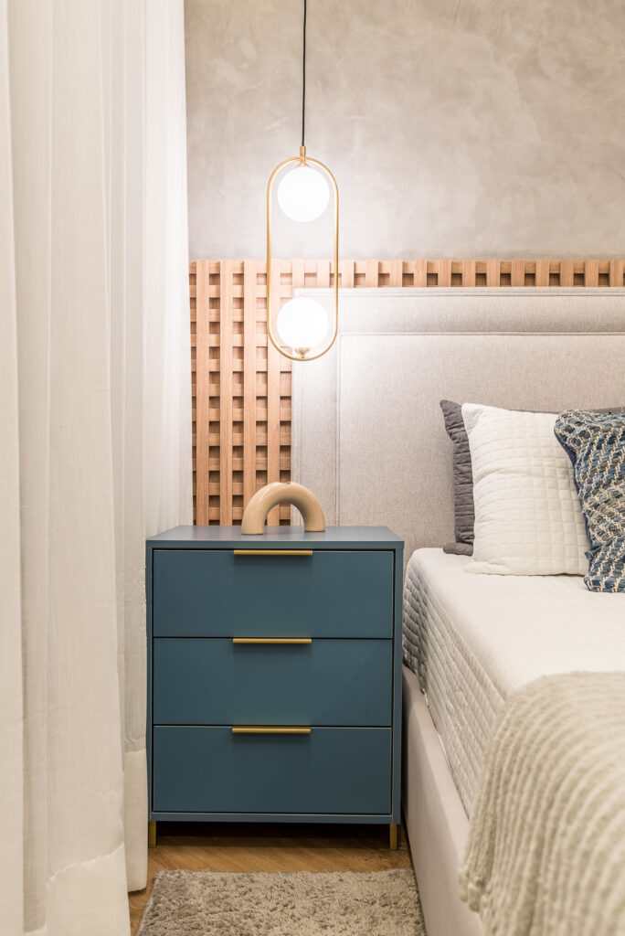 Quarto com painel amadeirado de treliça atrás da cabeceira, pendente ao lado da cama e mesa de cabeceira azul com gavetas