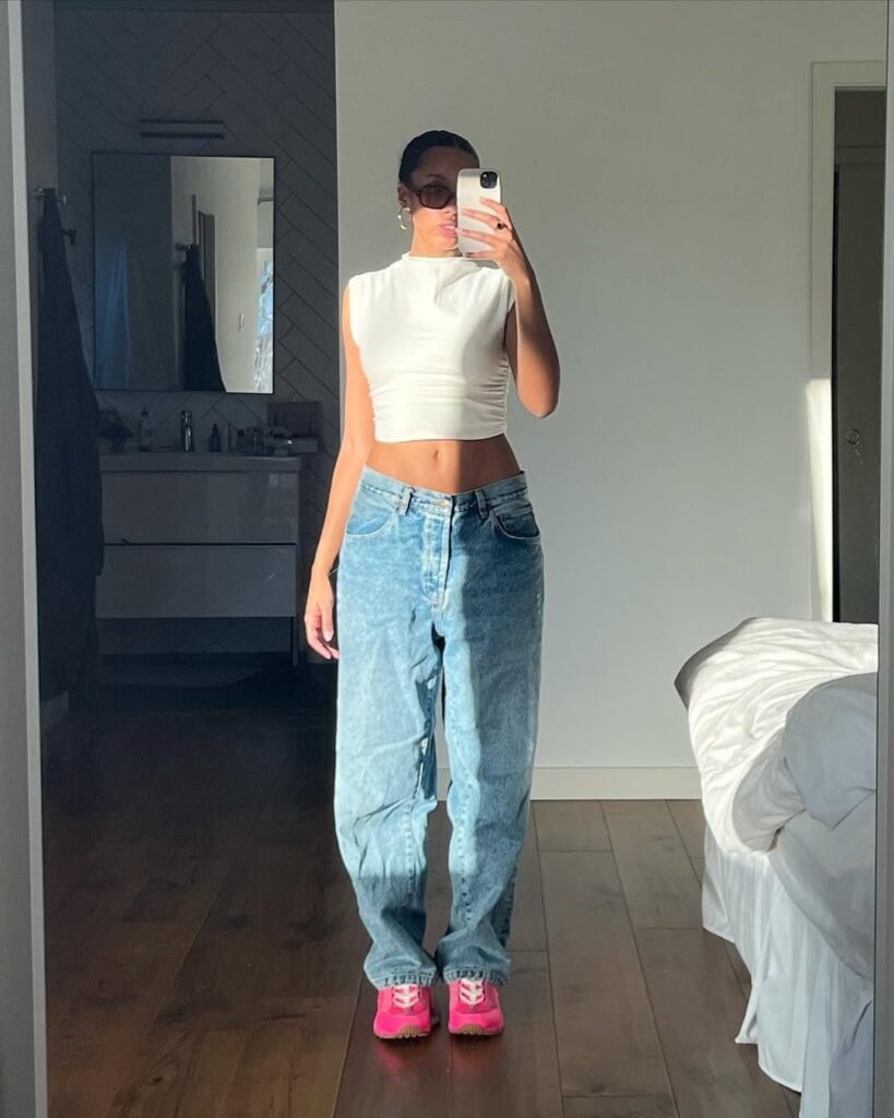 Mulher jovem e magra. Ela usa blusa branca, calça jeans e tênis rosa
