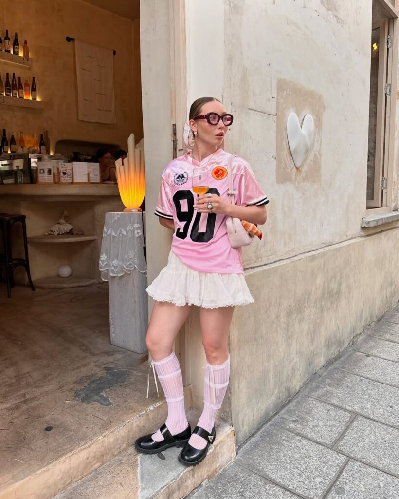 Mulher jovem, magra e de pele clara. Ela usa camiseta esportiva rosa, saia branca, sapato preto e meias brancas