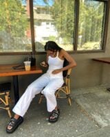 Mulher jovem, magra e de pele negra. Ela usa regata, calça jeans e papete.