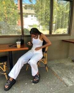 Mulher jovem, magra e de pele negra. Ela usa regata, calça jeans e papete.