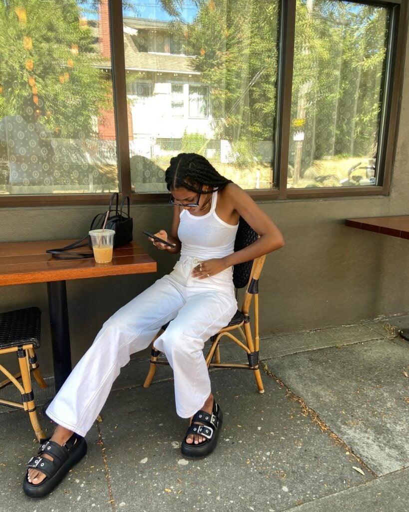 Mulher jovem, magra e de pele negra. Ela usa regata, calça jeans e papete.