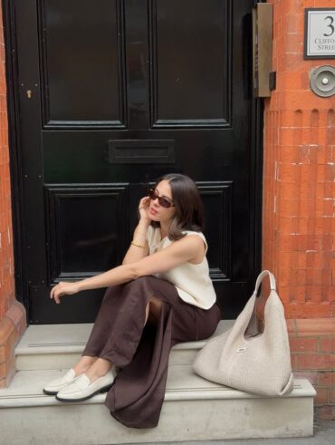 Tia Dewitt usando colete de tricô, saia midi marrom de cetim e loafer off-white