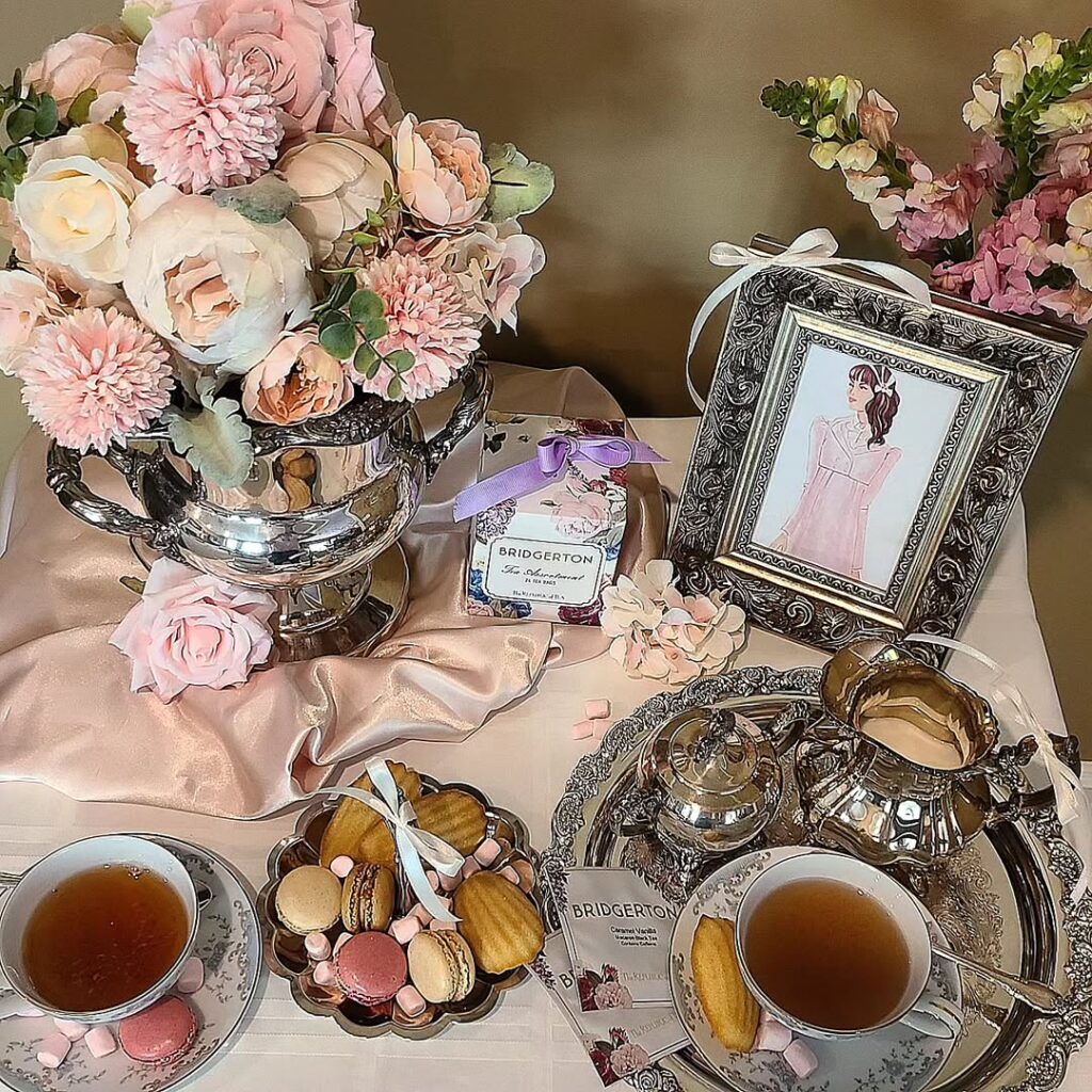 Mesa com toalha e flores rosa claro, xícaracas de chá, bandeja com macarons e madeleines