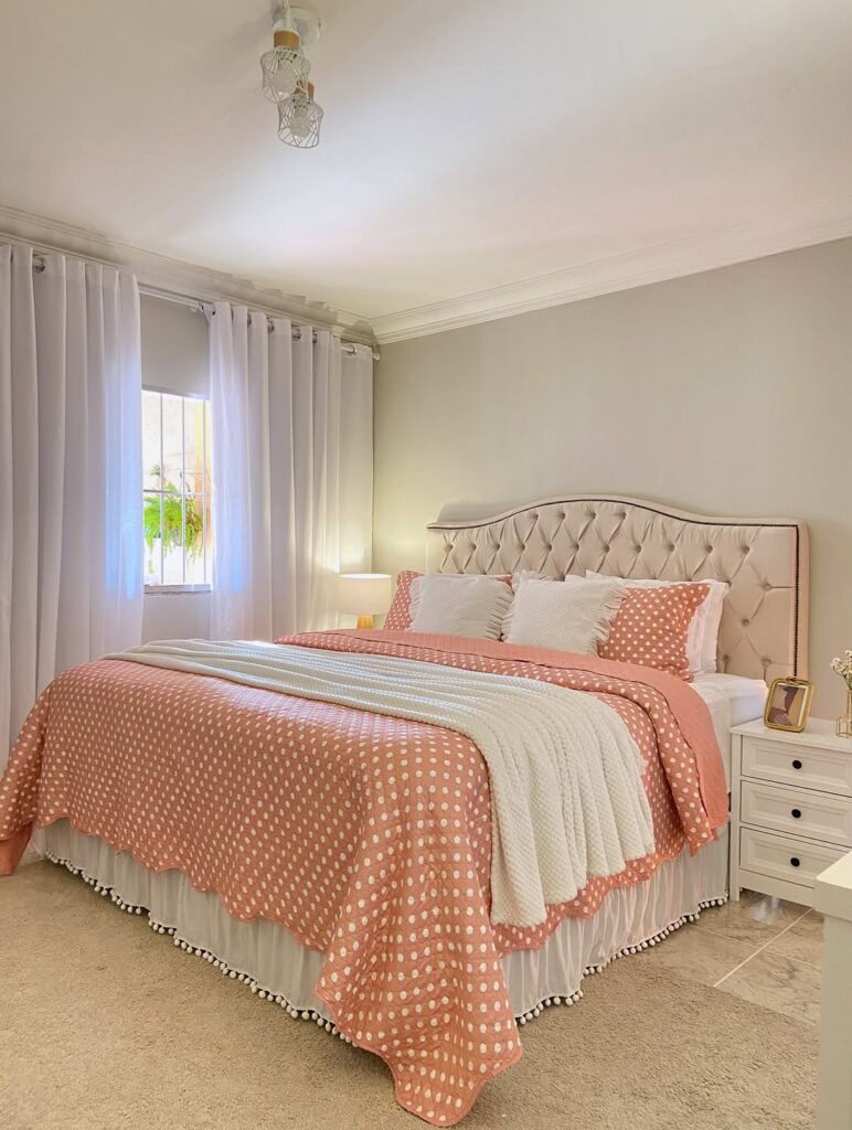 Cama com cabeceira capitonê bege, colcha rosa com bolinhas brancas, almofadas e cortina branca