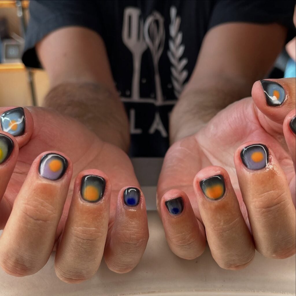 Unha curta com nail art maximalista em mão de pele negra com esmalte preto, laranja e azul