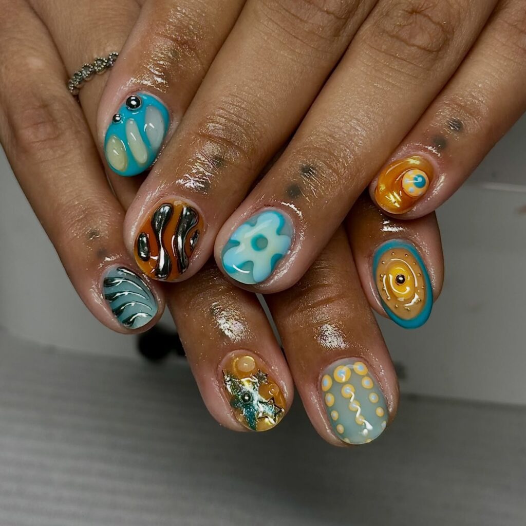Unhas curtas com nail art maximalista em mão de pele negra com esmalte azul, laranja, marrom e amarelo com metalizado e cromado