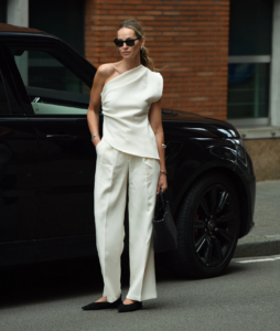 Mulher jovem magra e de pele clara com cabelos longos. Ela usa look moderno com blusa de um ombro só, calça e sapatilha