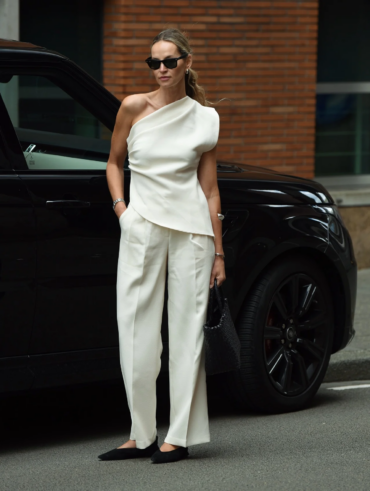 Mulher jovem magra e de pele clara com cabelos longos. Ela usa look moderno com blusa de um ombro só, calça e sapatilha