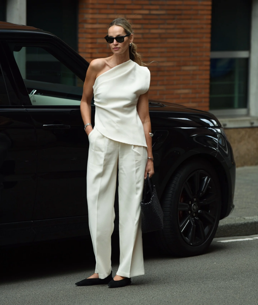 Mulher jovem magra e de pele clara com cabelos longos. Ela usa look moderno com blusa de um ombro só, calça e sapatilha