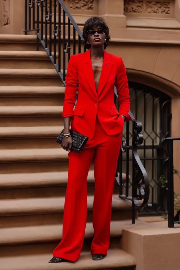 Mulher jovem, magra, alta e com pele negra. Ela tem cabelos curtos, franja e usa óculos. Ela veste blazer vermelho, calça vermelha e segura bolsa clutch