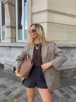 Mulher jovem de pele clara e cabelos loiros. Ela usa look elegante com blazer, blusa e bermuda e segura uma bolsa na mão direita.