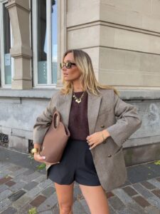 Mulher jovem de pele clara e cabelos loiros. Ela usa look elegante com blazer, blusa e bermuda e segura uma bolsa na mão direita.