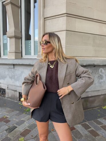 Mulher jovem de pele clara e cabelos loiros. Ela usa look elegante com blazer, blusa e bermuda e segura uma bolsa na mão direita.