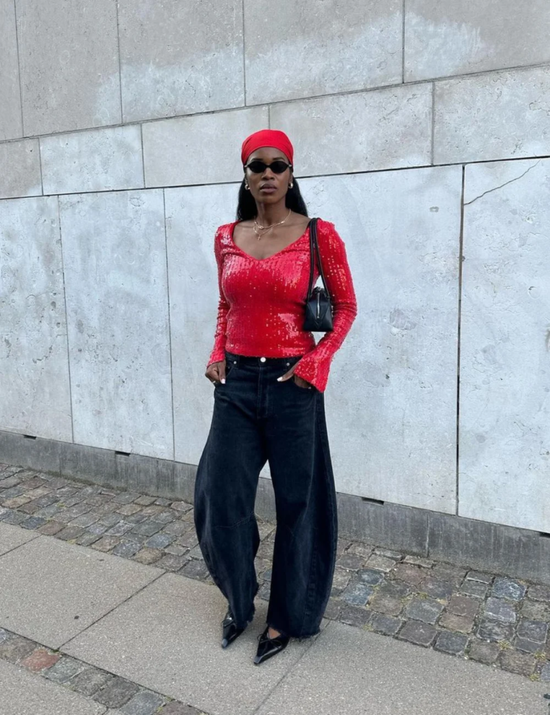 Mulher jovem, magra e de pele preta. Ela usa look com calça jeans barrel