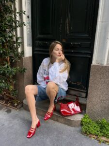 Mulher jovem, magra e com blusão e bermuda jeans. Ela calça sapato boneca vermelho e usa uma bolsa vermelha.