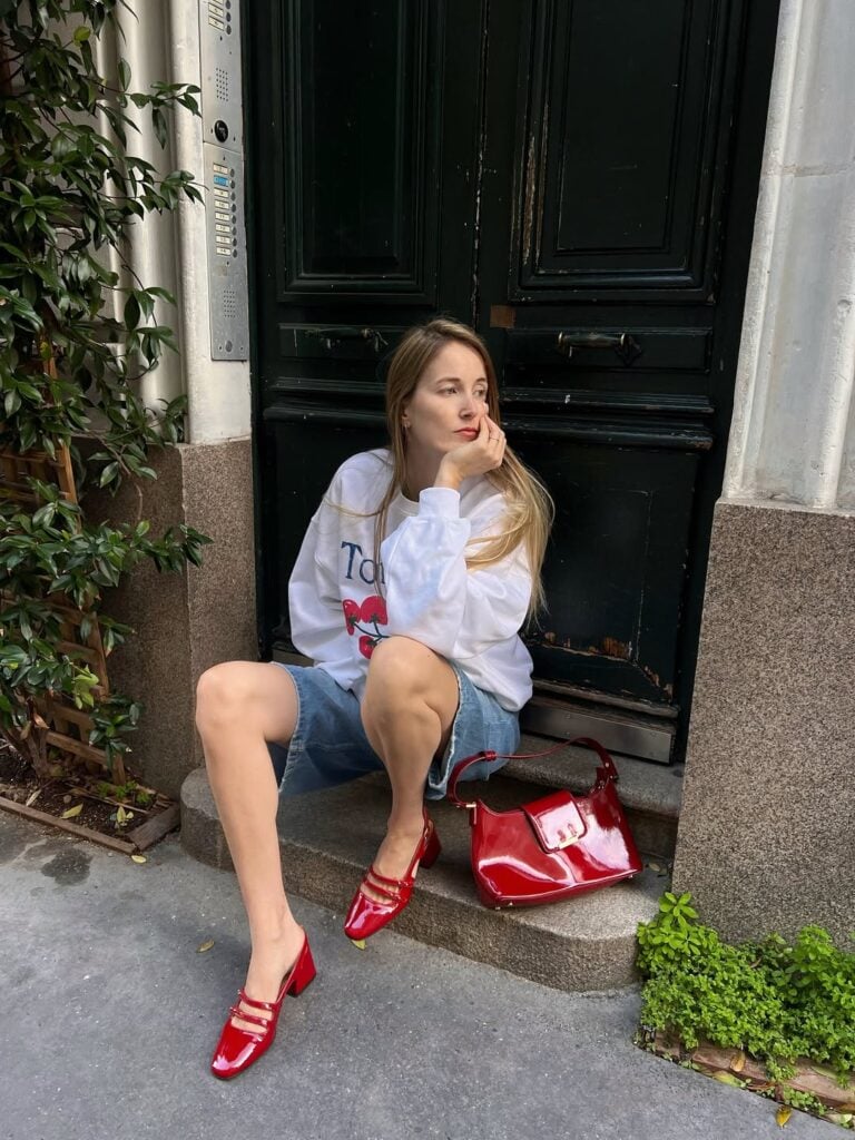 Mulher jovem, magra e com blusão e bermuda jeans. Ela calça sapato boneca vermelho e usa uma bolsa vermelha.