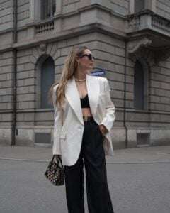 Mulher jovem, magra e alta com pele clara e cabelos loiros e longos. Ela usa blazer branco, top e calça preta e bolsa pequena estampada.