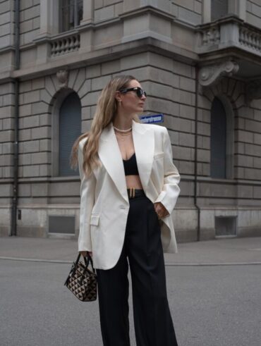 Mulher jovem, magra e alta com pele clara e cabelos loiros e longos. Ela usa blazer branco, top e calça preta e bolsa pequena estampada.