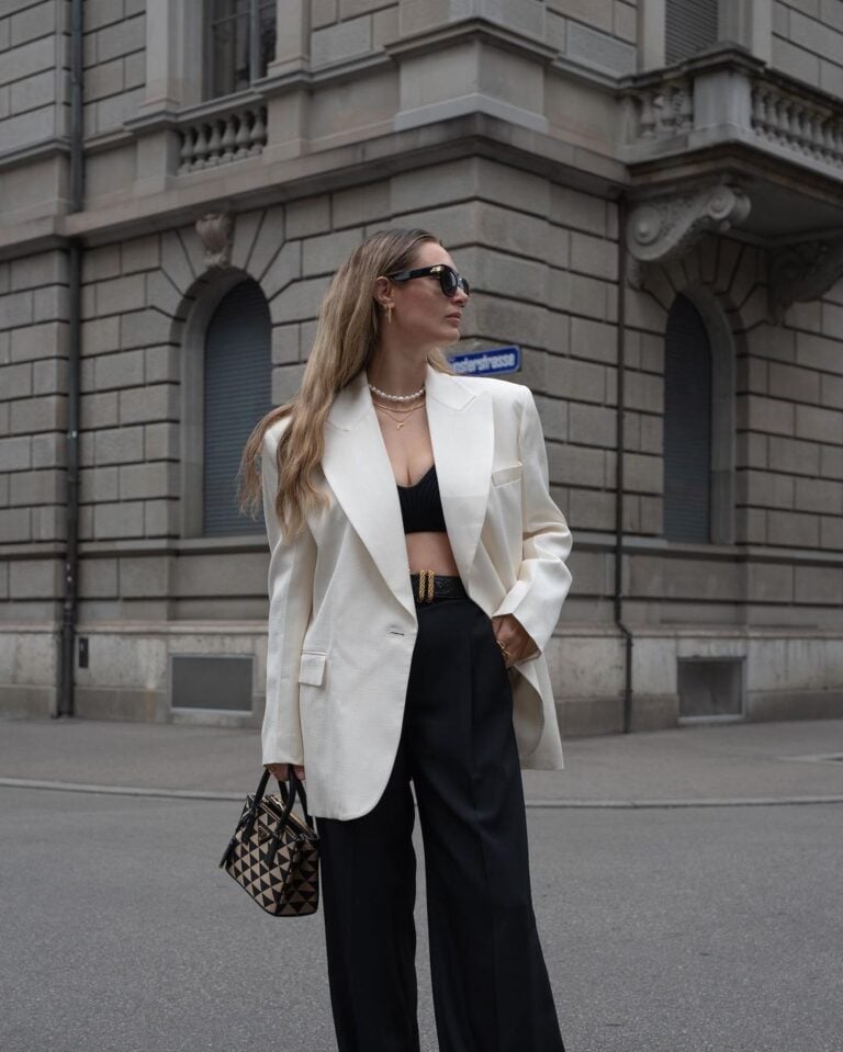 Mulher jovem, magra e alta com pele clara e cabelos loiros e longos. Ela usa blazer branco, top e calça preta e bolsa pequena estampada.
