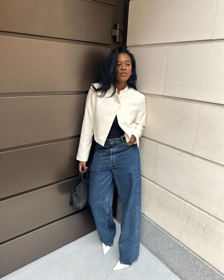 Mulher jovem, magra e de pele negra. Ela usa blazer branco e calça jeans