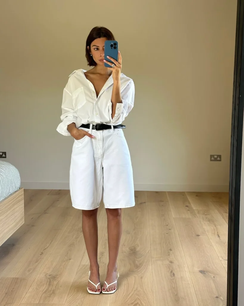 Mulher jovem de pele clara e cabelo curto. Ela usa camisa branca de manga longa, bermuda jeans com cinto preto e chinelo kitten heels.