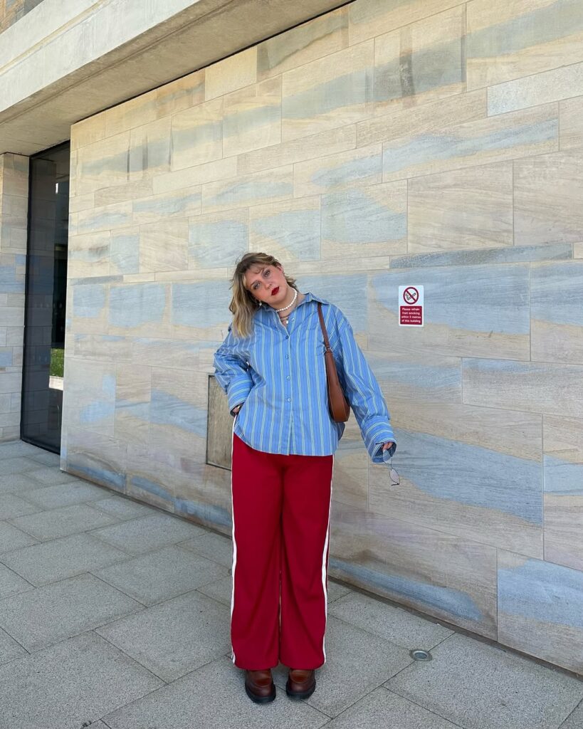 Mulher jovem, mid size e com pele clara. Ela veste camisa de manga longa azul com listras brancas, calça vermelha com listras brancas estilo esportivo e botas na cor marrom.
