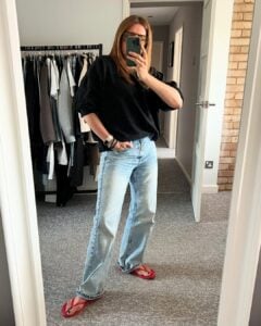 Mulher jovem, magra e de pele clara. Ela usa blusão preto, calça jeans e chinelo vermelho