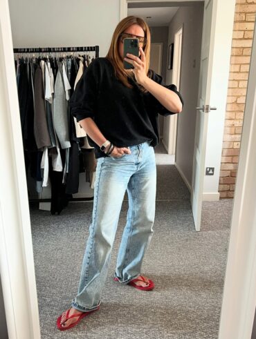 Mulher jovem, magra e de pele clara. Ela usa blusão preto, calça jeans e chinelo vermelho