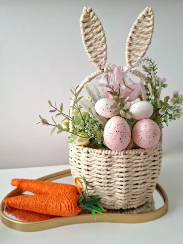 Cesto de palha com orelhas de coelho, ovinhos coloridos, cenouras feitas com linhas coloridas em bandeja espelhada