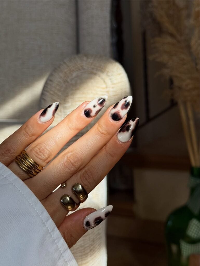 Mão de pele clara, com anéis em dourado e unha amendoada com estampa de vaca