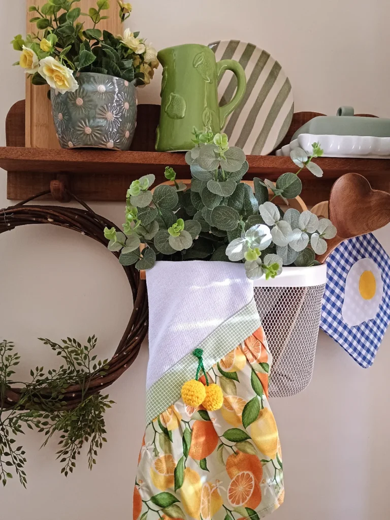 Prateleira de madeira com plantas, bule verde e pano de prato com estampa de laranjas