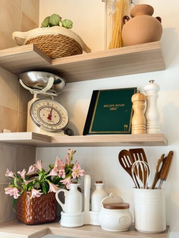 Cozinha com prateleiras no canto, balança, livro de receitas e utensílios de cozinha