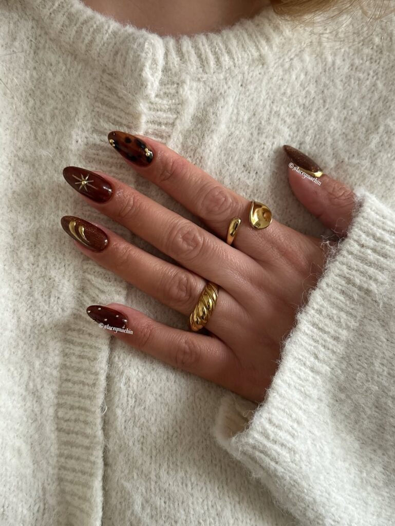Mão de pele clara, com anéis em dourado e unha marrom decorada