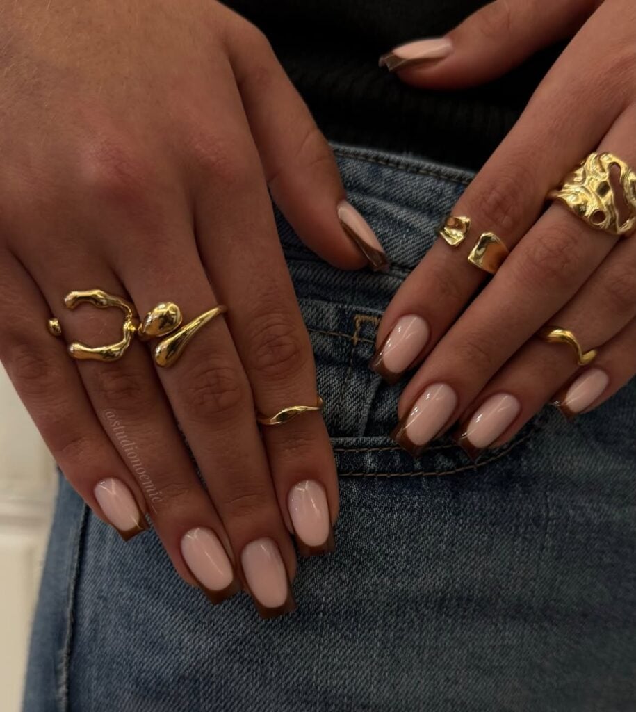 Mãos de pele clara, com anéis em dourado e unha quadrada neutra com francesinha marrom