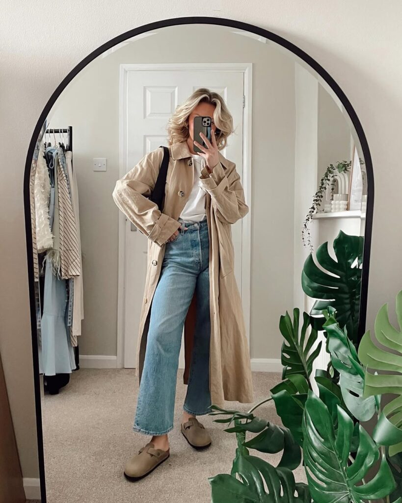Mulher jovem, magra e de pele clara. Ela usa trench coat, blusa, calça jeans e clogs