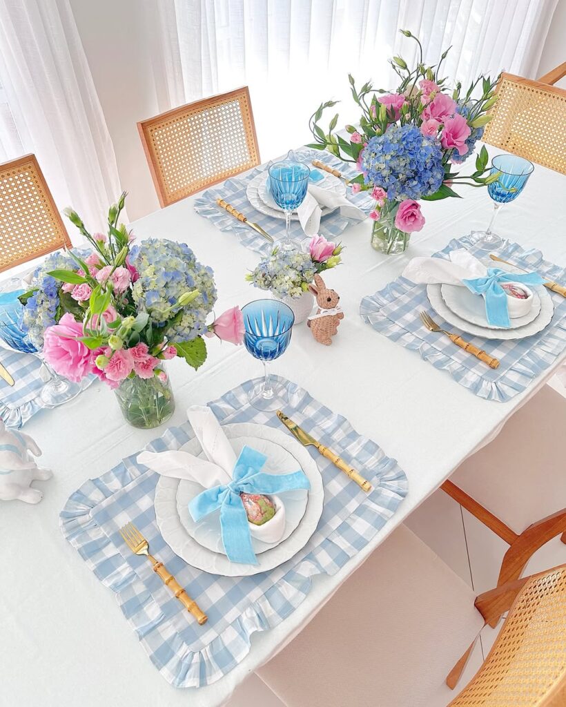 Mesa de Páscoa com jogos americanos xadrez em azul e branco, arranjos com flores rosas e azuis, e guardanapos em formato de orelhinhas