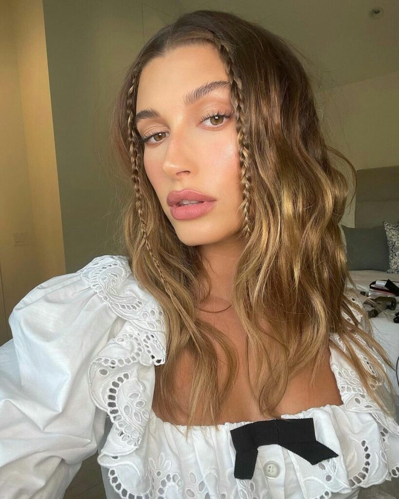 Hailey Bieber com blusa branca e cabelo solto com duas trancinhas na frente