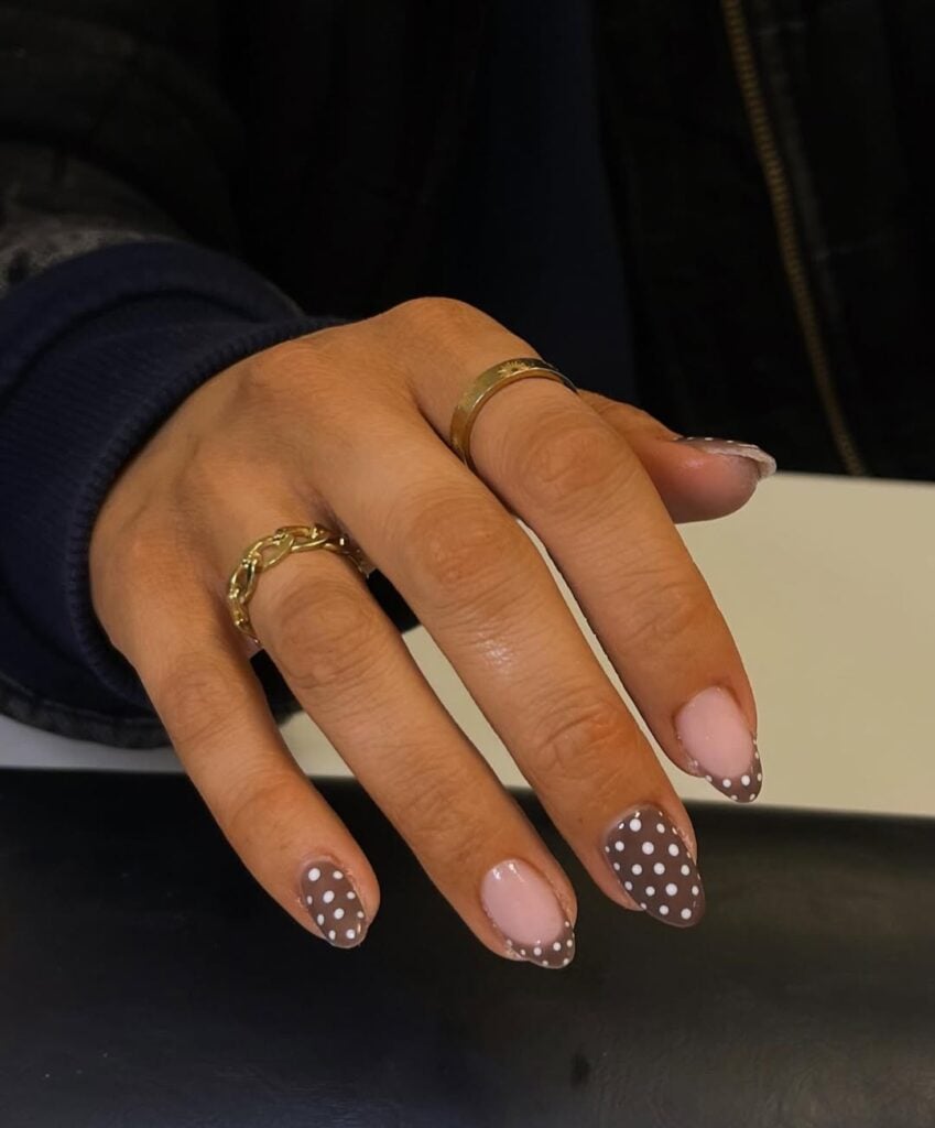 Mão de pele clara, com anéis em dourado e unha amendoada marrom com decoração de poá