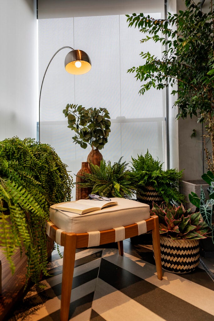 Ambiente com muitas plantas, luminária de piso e banqueta com livro e óculos
