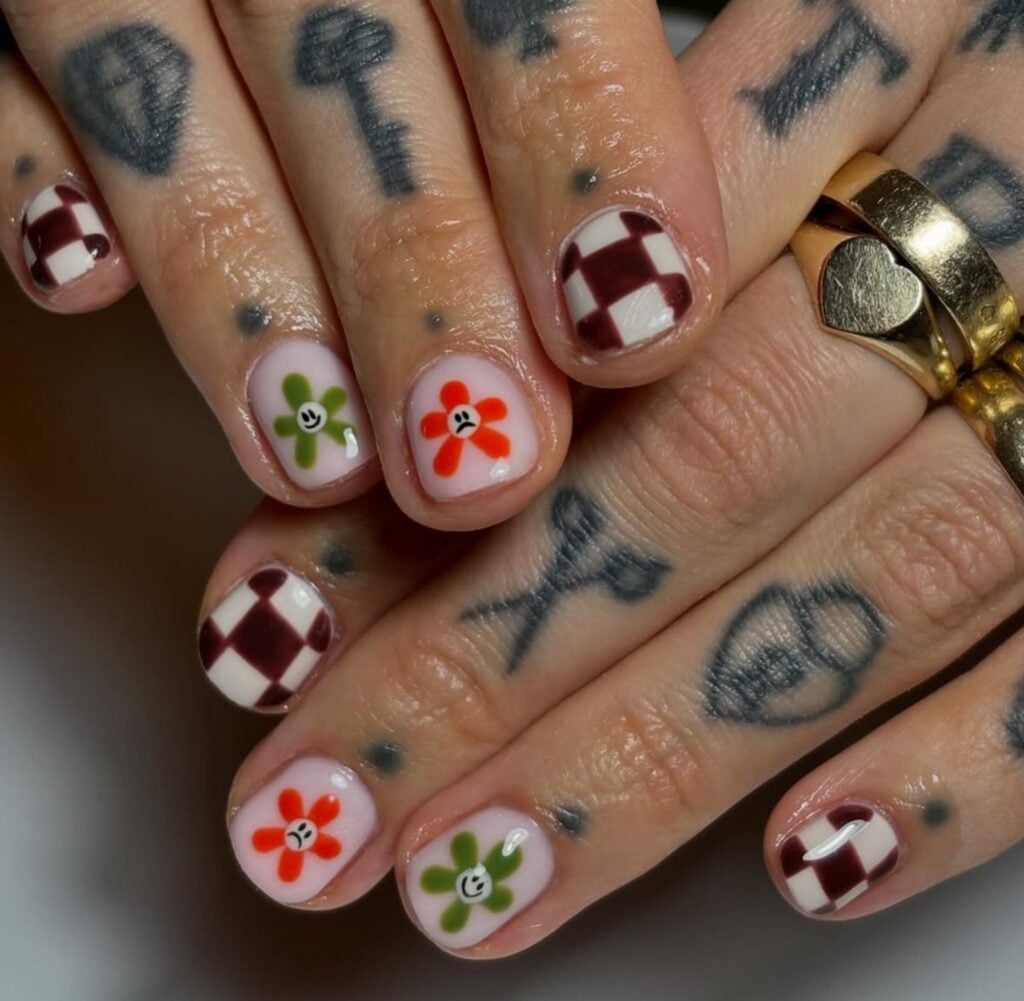 Mão de pele branca com tatuagem e unha com flor e emojis, além de unha checkerboard nails ou unhas de tabuleiro de xadrez com efeito quadriculado
