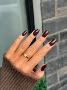 Mão de pele clara, com anel em dourado e unha amendoada marrom-escuro