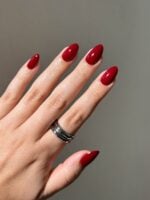 Mão de pele clara, com anel prata e unha amendoada vermelho-cereja