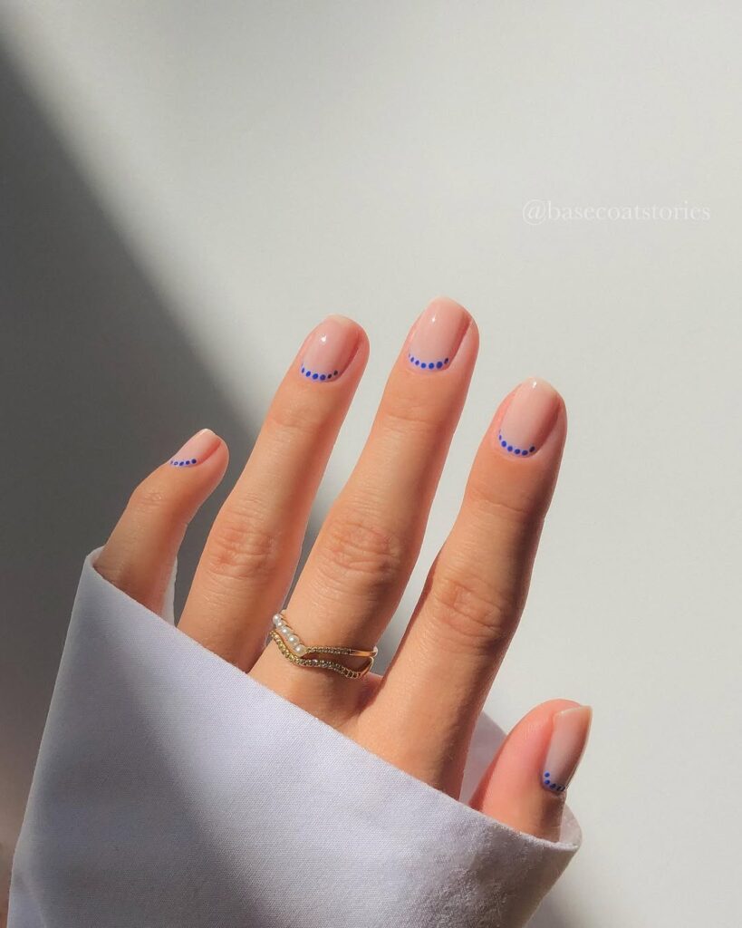 Mãos de pele clara, com anel dourado e unha neutra com francesinha invertida azul com bolinhas