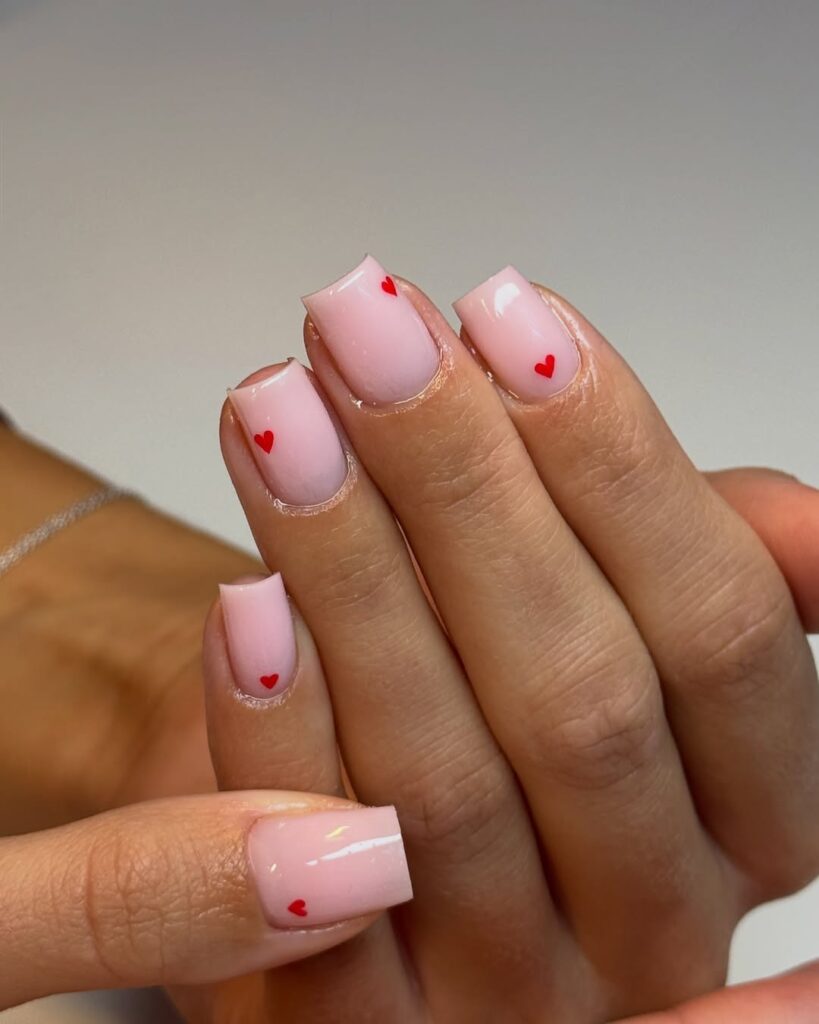 Mão de pele clara, com unha curta e quadrada rosa-claro com corações em vermelho 